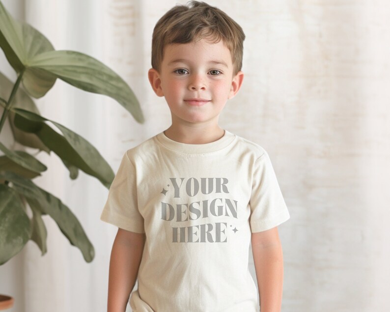 以下が含まれることがあります： 白いTシャツを着た小さな子供が、前面に「YOUR DESIGN HERE」という文字が印刷されています。