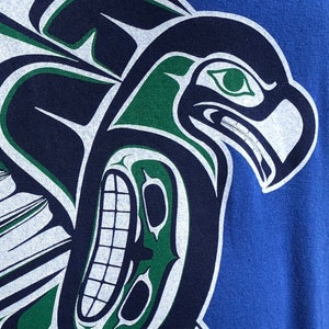 May include: A blue t-shirt with a green, white, and black stylized eagle design. The eagle has a large beak and a fierce expression.