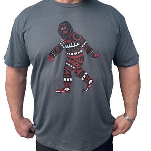 May include: A dark gray t-shirt with a red, black, and white graphic of a stylized Bigfoot walking. The graphic is in a Native American style.