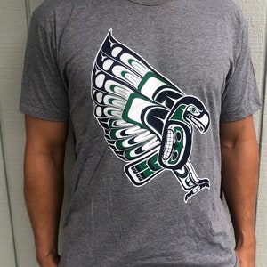 May include: A heather grey t-shirt with a green and blue stylized eagle design. The eagle is in a flying pose with its wings spread out.