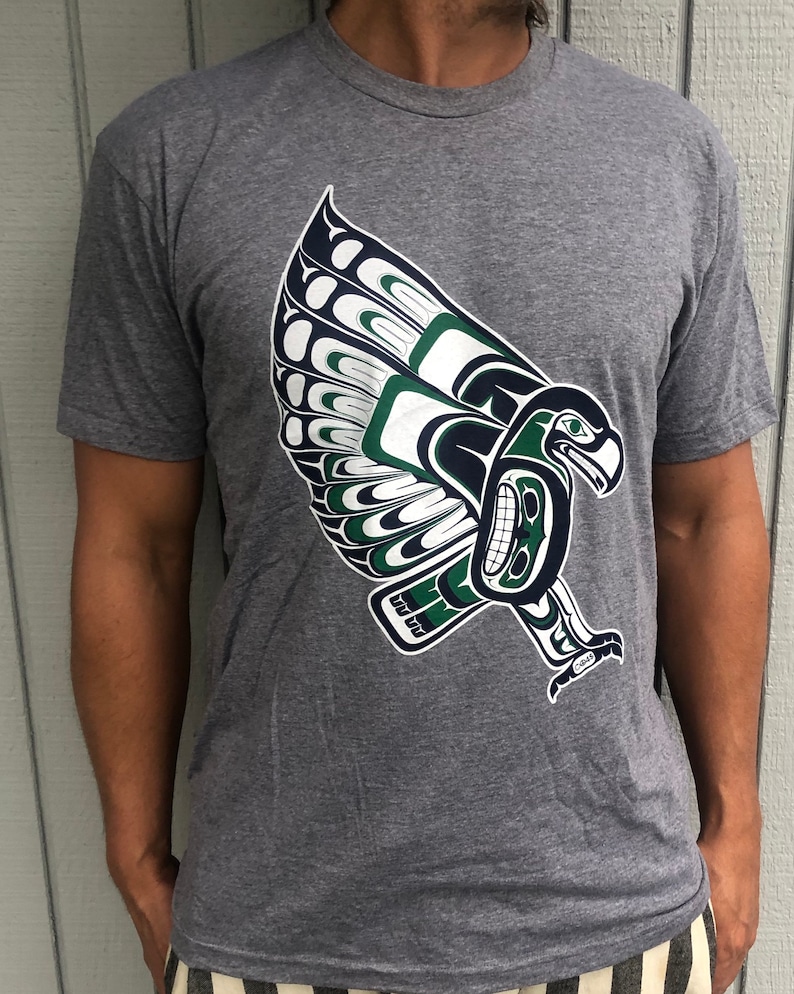 May include: A heather grey t-shirt with a green and blue stylized eagle design. The eagle is in a flying pose with its wings spread out.