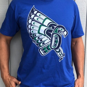 May include: A royal blue t-shirt with a green and white stylized eagle design. The eagle is in flight with its wings spread wide.