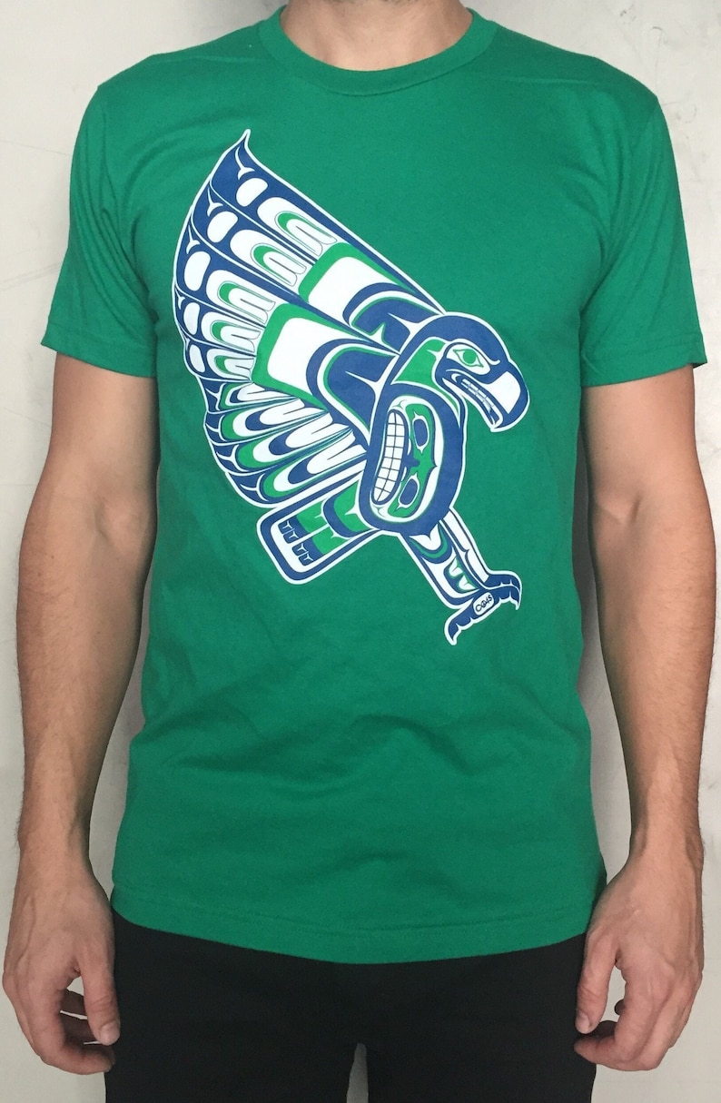 May include: A green t-shirt with a blue and green stylized eagle design. The eagle is in a flying pose with its wings spread out.