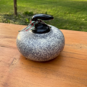 May include: A speckled, round ceramic object with a black top and a decorative ribbon. The object is gray with dark blue speckles and sits on a wooden surface. The ribbon has red, green, and white stripes.