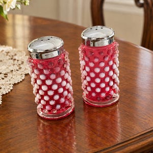 May include: A pair of cylindrical salt and pepper shakers featuring a vibrant red glass body with a raised white hobnail pattern. Each shaker is topped with a polished silver-coloured metal cap with small dispensing holes on the top surface.