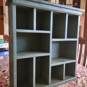 May include: A sage green wooden wall shelf featuring a rectangular frame with multiple compartments of varying sizes. The wood has a distressed finish with visible wear along the edges and surfaces, creating a textured appearance throughout.
