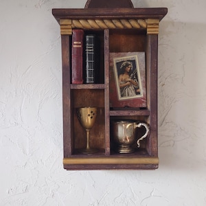 May include: A dark wood wall shelf with a rope-carved top edge, featuring four compartments. It holds two books labeled Nouveau Testament, a framed portrait of a person, a gold-toned metal chalice with cutouts, and a silver-toned metal handled cup.