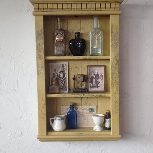 May include: A distressed yellow wall shelf with various vintage items. The shelf holds glass bottles, antique photos, a small clock, a teacup, and a blue bottle. A pink rose sits atop the shelf, adding a touch of color to the display.