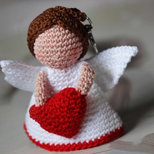 May include: A crochet angel keychain featuring a brown yarn head, a white dress with a red bottom border, white wings, and a red heart held in its arms. The figure has a light peach-colored face and is attached to a silver metal keyring loop.