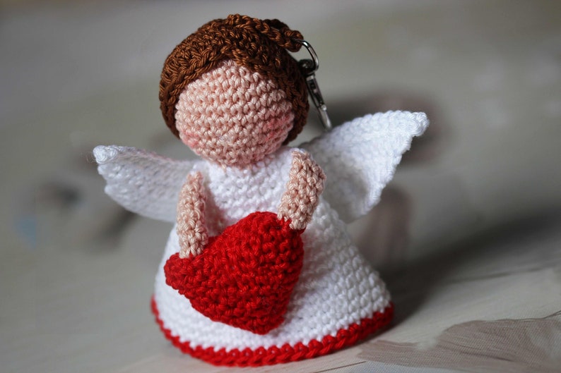 May include: A crochet angel keychain featuring a brown yarn head, a white dress with a red bottom border, white wings, and a red heart held in its arms. The figure has a light peach-colored face and is attached to a silver metal keyring loop.