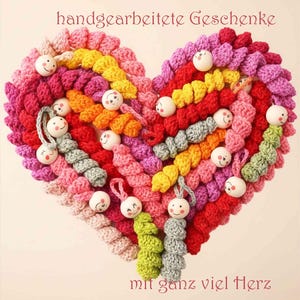 Könnte beinhalten: Ein handgefertigtes, herzförmiges Objekt aus farbenfrohem Garn in Rosa, Rot, Gelb und Grün. Verziert mit lächelnden Holzperlen, mit den Texten "handgearbeitete Geschenke" und "mit ganz viel Herz".