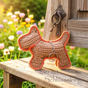 May include: A brown crocheted dog-shaped keychain with bright orange trim along the edges. The figure is attached to a silver-colored metal chain and a circular split ring. The text "hier mit Schlüsselring" is visible in the lower portion.