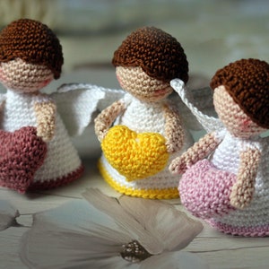 May include: Three small crocheted angel figurines with brown hair, white dresses, and white wings. Each angel holds a heart in its hands: one holds a dusty rose heart, one holds a bright yellow heart, and one holds a light pink heart.