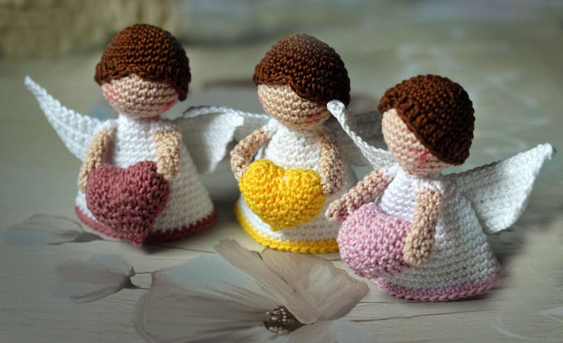 May include: Three small crocheted angel figurines with brown hair, white dresses, and white wings. Each angel holds a heart in its hands: one holds a dusty rose heart, one holds a bright yellow heart, and one holds a light pink heart.