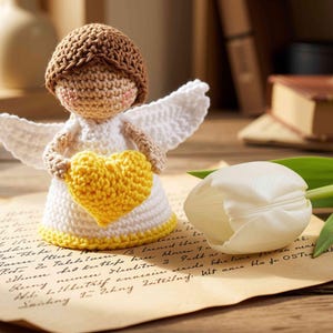 May include: A crocheted angel figurine with brown hair, white wings, and a white dress with a yellow hem, holding a yellow crocheted heart. The angel is positioned on a piece of aged paper with cursive script next to a single white tulip flower.