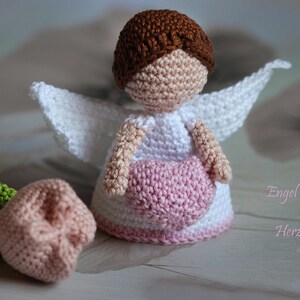 May include: A crocheted angel figurine with brown hair, a white dress, and white wings, holding a pink heart. Beside it sits a small crocheted flower with a pink bloom and green stem. The text "Engel mit Herz" is visible on the right side.