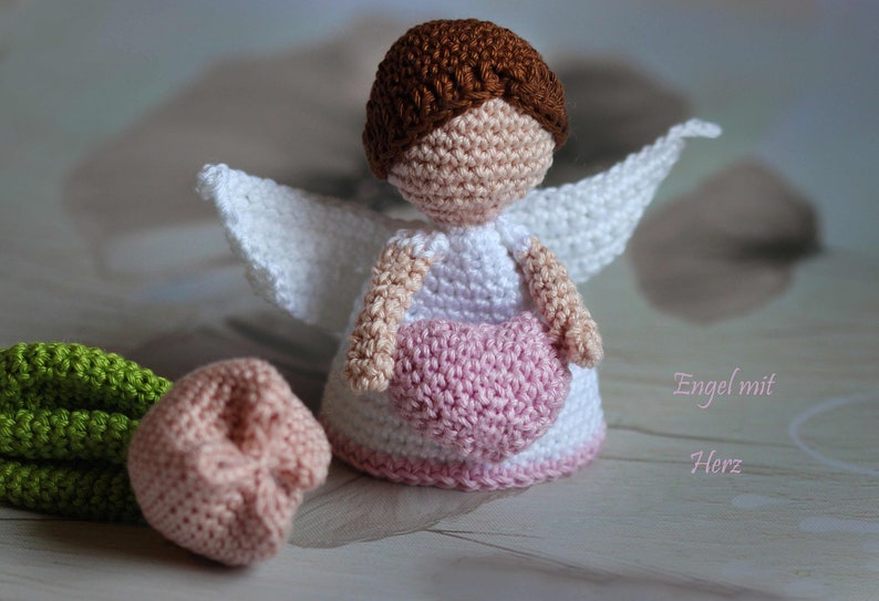 May include: A crocheted angel figurine with brown hair, a white dress, and white wings, holding a pink heart. Beside it sits a small crocheted flower with a pink bloom and green stem. The text "Engel mit Herz" is visible on the right side.