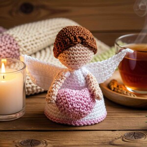 May include: A handcrafted crochet angel doll featuring a brown yarn head, white wings, and a light pink heart held in its arms. The doll has a white dress base with a subtle pink trim at the bottom, constructed from textured yarn stitches.