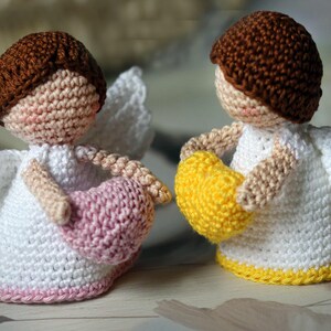 May include: Two crocheted angel figurines with brown hair, white dresses, and white wings. One angel holds a small pink heart, while the other holds a small yellow heart. Both figures have light skin tones and a simple, rounded, hand-crafted appearance.