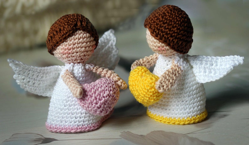 May include: Two crocheted angel figurines with brown hair, white dresses, and white wings. One angel holds a small pink heart, while the other holds a small yellow heart. Both figures have light skin tones and a simple, rounded, hand-crafted appearance.
