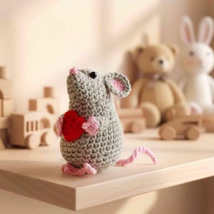 May include: A handmade crochet mouse in gray holding a red heart. The mouse has pink accents on its ears, feet, and tail. Other toys, including a teddy bear and a bunny, are in the background on a wooden shelf. The toys are in a neutral color palette.