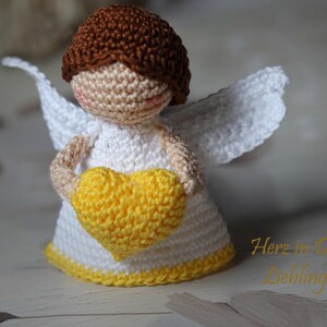 May include: A small crocheted angel figurine with brown hair, a white dress, and white wings. The angel holds a bright yellow crocheted heart in its hands. The text "Herz in Deiner Lieblingsfarbe" is visible in the lower right corner of the image.