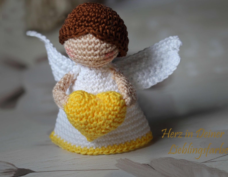 May include: A small crocheted angel figurine with brown hair, a white dress, and white wings. The angel holds a bright yellow crocheted heart in its hands. The text "Herz in Deiner Lieblingsfarbe" is visible in the lower right corner of the image.