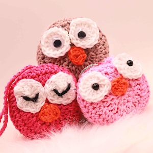 May include: Three crocheted owl-shaped items in shades of pink and brown. Each owl has large white eyes with black pupils and an orange beak. One owl has a loop for hanging. The owls are arranged on a soft, light-colored surface.