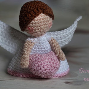 May include: A crocheted angel doll with brown hair, a white dress, and white wings, holding a pink heart. The doll has a beige face with pink cheeks. The text "Dein Engel mit Herz" is printed in pink in the lower right corner of the image.