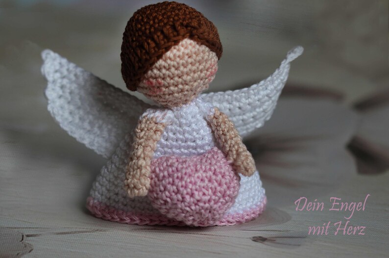 May include: A crocheted angel doll with brown hair, a white dress, and white wings, holding a pink heart. The doll has a beige face with pink cheeks. The text "Dein Engel mit Herz" is printed in pink in the lower right corner of the image.