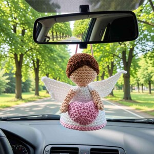 May include: A crocheted angel ornament hanging from a rearview mirror. The figure has brown hair, a tan face, white wings, and a white dress with a pink heart centered on the front. The ornament is suspended by a thin pink string in a vehicle.