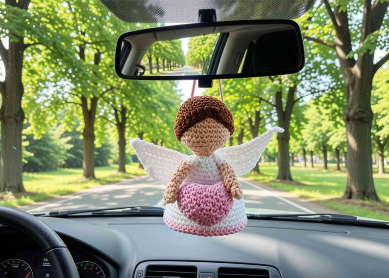 May include: A crocheted angel ornament hanging from a rearview mirror. The figure has brown hair, a tan face, white wings, and a white dress with a pink heart centered on the front. The ornament is suspended by a thin pink string in a vehicle.
