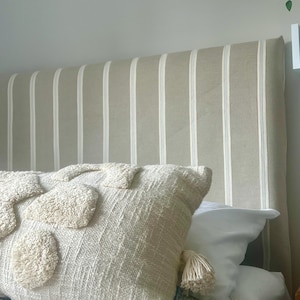 May include: A beige and white striped headboard with a textured, cream-colored pillow featuring tufted accents and a tassel. The bed is covered with a light blue blanket and white sheets, creating a cozy bedroom setting.