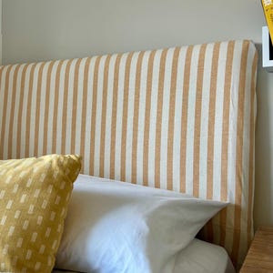Peut inclure: Un lit avec une tête de lit à rayures verticales beiges et blanches. Le lit est fait avec des draps blancs, un oreiller jaune à motifs et une couverture en tricot grise. Un livre intitulé "Yellowface" est visible.