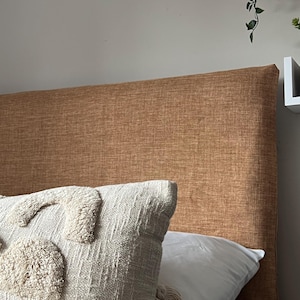 May include: A close-up of a brown upholstered headboard with a white and cream textured throw pillow on top. The headboard is made of a textured fabric and has a simple, modern design.