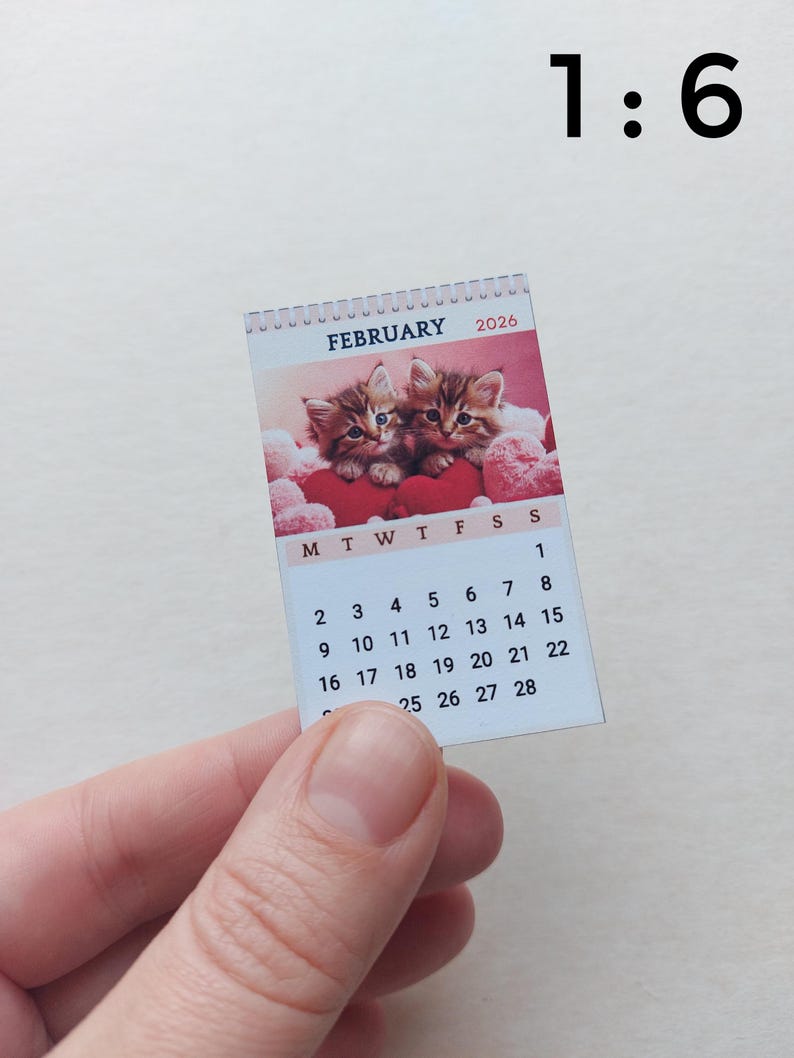 May include: A miniature 1:6 scale wall calendar for February 2026 featuring a photograph of two brown tabby kittens nestled among red heart-shaped cushions. The calendar displays the month, year, and a grid of dates from 1 through 28.