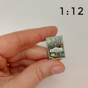 Puede incluir: Un libro en miniatura titulado "Enciclopedia de plantas acu&aacute;ticas" con ilustraciones de nen&uacute;fares y otras plantas acu&aacute;ticas. El libro est&aacute; a escala 1:12.