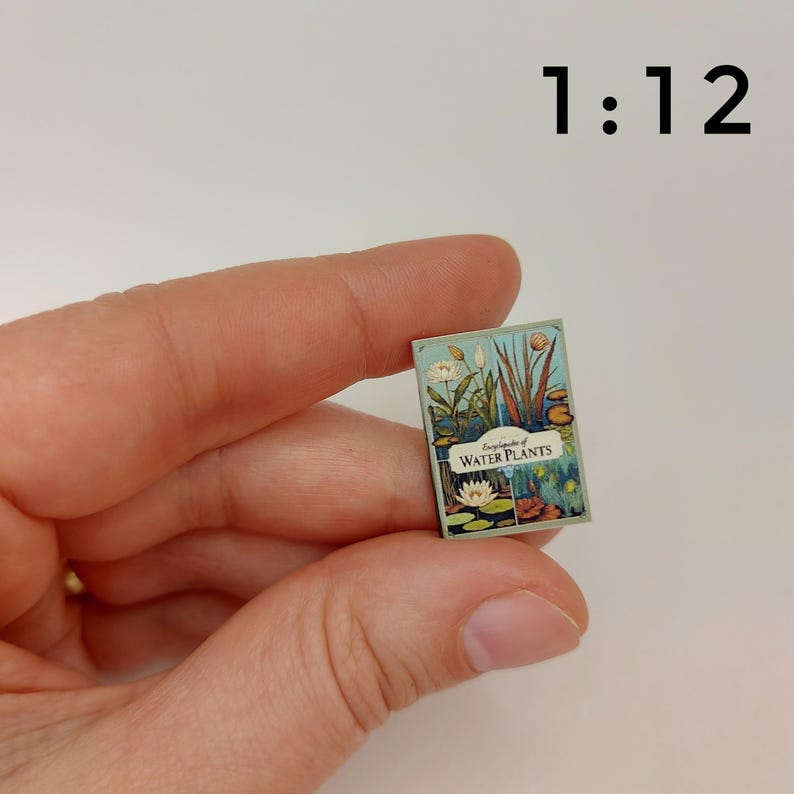 Puede incluir: Un libro en miniatura titulado "Enciclopedia de plantas acu&aacute;ticas" con ilustraciones de nen&uacute;fares y otras plantas acu&aacute;ticas. El libro est&aacute; a escala 1:12.
