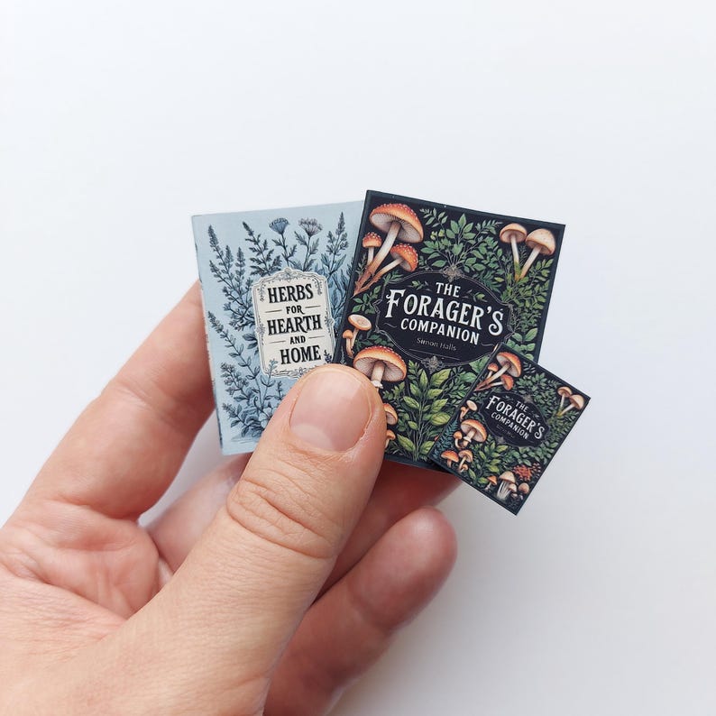 Puede incluir: Libros en miniatura con ilustraciones bot&aacute;nicas. Uno es azul claro con el texto "Herbs for Hearth and Home". Los otros son oscuros con ilustraciones de setas y el texto "The Forager's Companion".