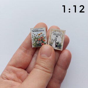 Puede incluir: Dos libros en miniatura, probablemente para una casa de mu&ntilde;ecas, se sostienen en una mano. Un libro se titula "Illustrated Guide Wildflowers" y presenta una ilustraci&oacute;n floral. El otro libro se titula "The Lady's Herbarium" con una mujer cuidando plantas.