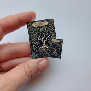 Puede incluir: Dos libros en miniatura con el t&iacute;tulo "MATERIA HERBARIA" se sostienen en una mano. Los libros tienen una cubierta azul oscuro con un dise&ntilde;o de &aacute;rbol dorado. El &aacute;rbol tiene ra&iacute;ces, tronco, ramas y hojas.