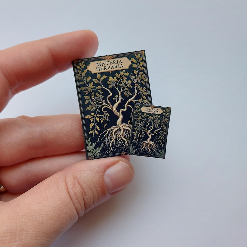 Puede incluir: Dos libros en miniatura con el t&iacute;tulo "MATERIA HERBARIA" se sostienen en una mano. Los libros tienen una cubierta azul oscuro con un dise&ntilde;o de &aacute;rbol dorado. El &aacute;rbol tiene ra&iacute;ces, tronco, ramas y hojas.