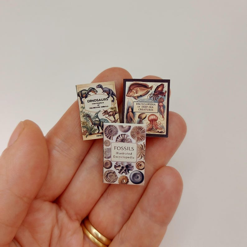 Puede incluir: Tres libros en miniatura con ilustraciones coloridas. Los libros se titulan "Dinosaurs", "Fossils Illustrated Encyclopedia" y "Encyclopedia of Deep-Sea Creatures".