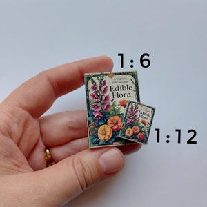 Puede incluir: Dos libros en miniatura con el t&iacute;tulo "Edible Flora" e ilustraciones florales. Los libros se sostienen en una mano. Las ilustraciones presentan flores rosas, naranjas y amarillas. El texto "Edible Flora" es visible.