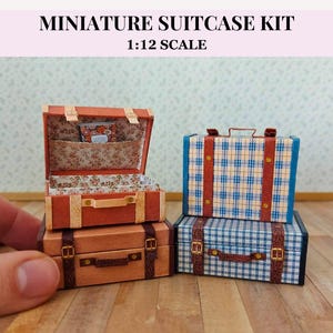 May include: Miniature suitcase kit in 1:12 scale. The image shows two stacks of suitcases, one open and one closed. The open suitcase is orange with a floral interior. The closed suitcases are brown and blue plaid with brown straps.