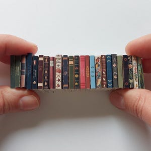 Puede incluir: Una colecci&oacute;n de libros en miniatura con diversas cubiertas de colores, dispuestos en fila y sostenidos entre dos manos.