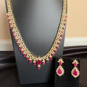 Puede incluir: Un conjunto de collar y pendientes dorados. El collar presenta un diseño en forma de hoja con piedras preciosas claras y rosas. Los pendientes tienen un diseño similar, con una piedra preciosa rosa en forma de lágrima en el centro. La joyería se exhibe en un soporte negro.