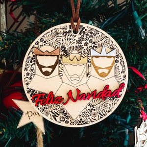 May include: Wooden Christmas ornament featuring three kings with crowns and beards, with the words "Feliz Navidad" in red. The ornament has a floral design around the edges and a brown leather cord for hanging.