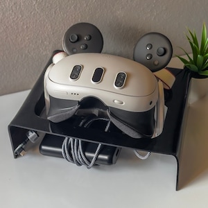Op de afbeelding: Een zwarte standaard met een witte VR-headset en twee zwarte controllers. De standaard heeft een groef voor de headset en een plek voor de controllers om te zitten. Er is een zwarte powerbank en een kabel op de standaard.