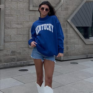 May include: A person wearing a blue sweatshirt with "KENTUCKY" in white lettering, denim shorts, and white cowboy boots. The sweatshirt has a mock neck. The person is wearing sunglasses and hoop earrings.